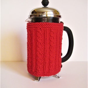 May include: A red knitted cozy for a French press coffee maker. The cozy is made with a cable knit pattern and has a silver metal base.