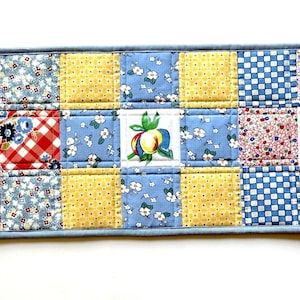 May include: A rectangular quilted table runner featuring a patchwork design of various floral, gingham, and fruit-patterned fabric squares in red, yellow, and blue. The runner includes three central squares depicting cherries, a peach, and blueberries.