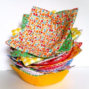 May include: A stack of colorful, quilted bowl cozies. The cozies feature various floral, gingham, and polka dot patterns in red, yellow, green, and blue. The bottom cozy is yellow with small polka dots.