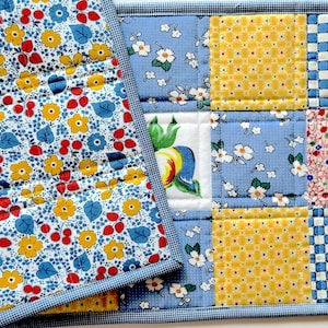 May include: A quilted fabric runner featuring a patchwork design of various floral and geometric patterns in yellow, blue, red, and white. The piece is finished with a blue and white checkered binding along the edges and visible grid stitching.