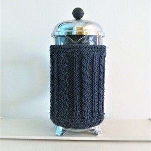 May include: A dark blue knitted cozy for a French press coffee maker. The cozy is made with a cable knit pattern and has a silver metal base.