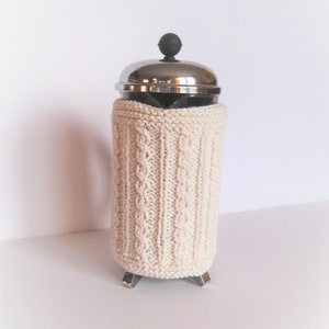 May include: A white knitted cozy for a French press coffee maker. The cozy has a cable knit pattern and is made of soft yarn.