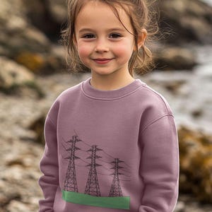 May include: A young person wearing a mauve sweatshirt with a graphic of three black power lines and towers on a green field.