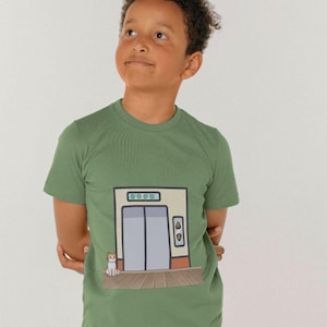 May include: A green t-shirt featuring a cartoon illustration of a lift with a cat sitting in front of it. The lift has a light brown frame and a grey door. The cat is white with orange markings.