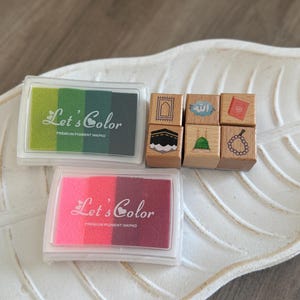 May include: A set of six wooden stamps with Islamic symbols, including a mosque, a prayer rug, a book, a hand holding prayer beads, and the Arabic word for God. The stamps are arranged in a row on a white surface. The set also includes two ink pads, one green and one pink, with the words "Let's Color" and "Premium Pigment Inkpad" printed on them.