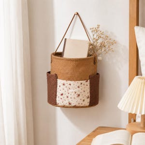 May include: A hanging wall storage basket made of brown burlap with a central cream-colored pocket featuring a small red floral pattern. The basket has two dark brown side pockets and is suspended by a thin brown cord from a wall hook.
