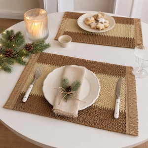 May include: Two rectangular woven placemats featuring alternating horizontal bands of dark brown and light beige with fringed edges. Each setting includes a white scalloped plate, a beige linen napkin, a fork, and a knife with white handles.