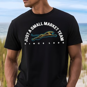 May include: Black t-shirt with the text "JUST A SMALL MARKET TEAM" in a semi-circle above a teal and gold jaguar logo. The text "SINCE 1995" is below the logo. The shirt is short-sleeved.