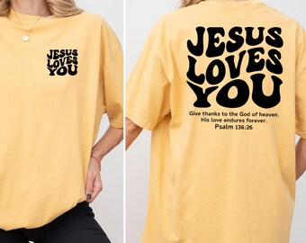 May include: Mustard yellow t-shirt with the text "JESUS LOVES YOU" in black on the front and back. The back of the shirt also includes the Bible verse Psalm 136:26. The shirt is a casual, oversized fit.
