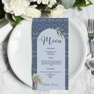 Puede incluir: Una tarjeta de menú con un borde de patrón geométrico azul y blanco, con la palabra "Menú" en escritura elegante. El menú enumera entrantes, platos principales y postres, con los nombres "Sarah & Omar" en la parte inferior. La tarjeta está sobre un plato blanco con detalles florales.