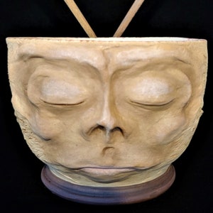 May include: A ceramic bowl shaped like a face with closed eyes. The bowl is resting on a wooden base and two wooden chopsticks are resting on the rim of the bowl.