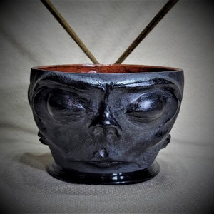 May include: Black ceramic bowl with a face sculpted on the front. The face has closed eyes, a prominent nose, and a slightly open mouth.
