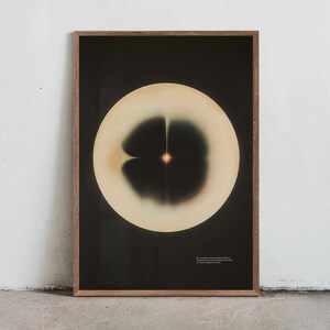 May include: A framed art print featuring an abstract design. The artwork showcases a large, off-white circular shape against a black background. Within the circle, a dark, symmetrical pattern is visible, with a central, glowing point. The frame is made of wood.