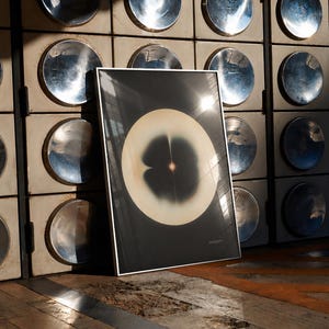 May include: A framed abstract art print featuring a large, light-colored circular shape with a dark center and a small, bright spot. The artwork is set against a dark background and leans against a wall with metallic circular accents.