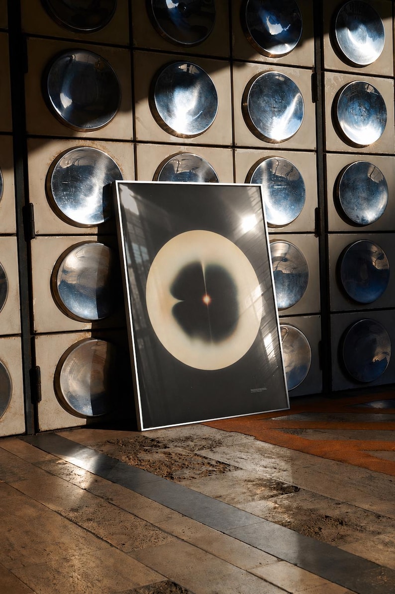 May include: A framed abstract art print featuring a large, light-colored circular shape with a dark center and a small, bright spot. The artwork is set against a dark background and leans against a wall with metallic circular accents.