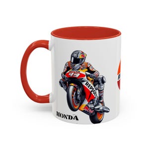 Peut inclure: Une tasse en céramique blanche avec une bordure et une poignée rouges. La tasse présente une illustration de dessin animé d'un pilote de moto sur une moto Honda rouge et blanche avec le logo Repsol. Le texte "HONDA" est imprimé au bas de la tasse.