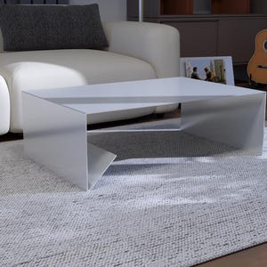 Peut inclure: Une table basse blanche moderne au design géométrique angulaire. La table est posée sur un tapis texturé gris clair, avec un canapé blanc et un coussin gris foncé en arrière-plan. Deux guitares sont visibles.