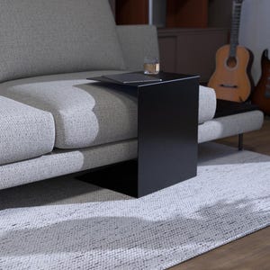 May include: A modern living room scene featuring a light gray sofa, a black side table, and a white rug. A glass sits on the table. Guitars and vinyl records are in the background, suggesting a music or entertainment theme.