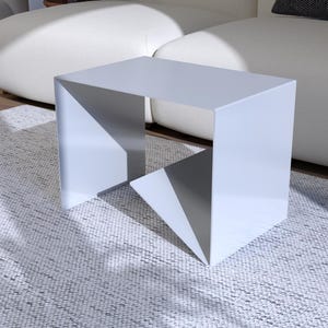Puede incluir: Una mesa auxiliar moderna y geométrica en color gris claro. La mesa tiene un diseño único con patas angulares y una parte superior rectangular. Se coloca sobre una alfombra gris claro, cerca de un sofá blanco. La mesa mide aproximadamente 46 cm de largo, 46 cm de ancho y 41 cm de alto.