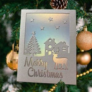 Op de afbeelding: Een zilveren kerstkaart met een uitgesneden ontwerp met een boom, huizen, een hert en sterren. De woorden "Merry Christmas" zijn onderaan uitgesneden. De kaart is versierd met gouden en groene kerstversieringen.