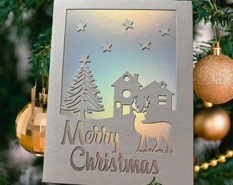 May include: A silver Christmas card with a cut-out design featuring a tree, houses, a deer, and stars. The words "Merry Christmas" are cut out at the bottom. The card is decorated with gold and green Christmas ornaments.