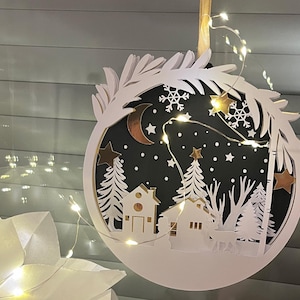 Puede incluir: Adorno navideño circular blanco y en capas con una escena invernal. El adorno presenta un fondo negro con una luna creciente, estrellas, copos de nieve, casas, árboles y un ciervo. Una cadena de luces rodea el adorno.
