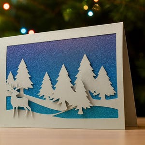 May include: A handmade Christmas card featuring a winter scene with a deer and pine trees cut from white paper. The background has a gradient of blue and teal glitter. The card is displayed in front of a Christmas tree.