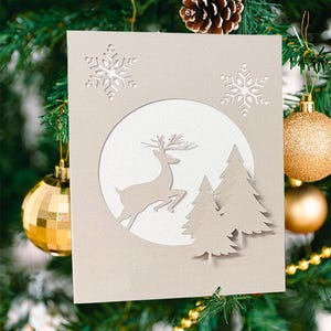以下が含まれることがあります： ベージュ色のクリスマスカード。白い円の中に、2本の松の木の前にジャンプするトナカイが描かれた切り絵のデザイン。円の上には2つの雪の結晶の切り抜きがあります。カードは金のオーナメントと松の葉で飾られています。