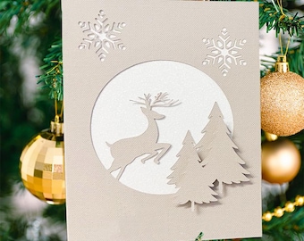May include: A beige Christmas card with a cut-out design featuring a reindeer leaping in front of two pine trees, set within a white circle. Two snowflake cutouts are above the circle. The card is decorated with gold ornaments and pine needles.