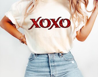 May include: Cream-colored t-shirt with the word "XOXO" in red glitter lettering. The short-sleeved shirt is paired with light-wash blue jeans. The shirt is a casual, everyday wear item.