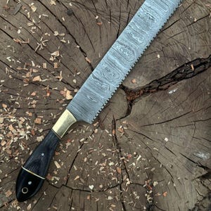 May include: A handcrafted bread knife with a patterned steel blade and a serrated edge. The handle is black with brass accents and a rounded end. The knife is displayed on a rustic wooden surface.