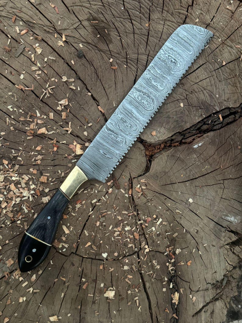 May include: A handcrafted bread knife with a patterned steel blade and a serrated edge. The handle is black with brass accents and a rounded end. The knife is displayed on a rustic wooden surface.