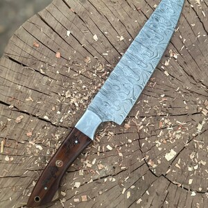 May include: A Damascus steel chef's knife with a dark wood handle. The blade has a unique swirling pattern. The knife is resting on a weathered wooden surface, showcasing the craftsmanship and design of the kitchen tool.