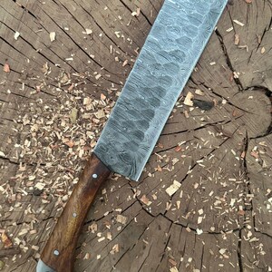 May include: A large, rectangular cleaver knife with a patterned steel blade and a wooden handle. The handle is secured with rivets and has a metal end cap. The knife rests on a wooden surface with wood shavings.