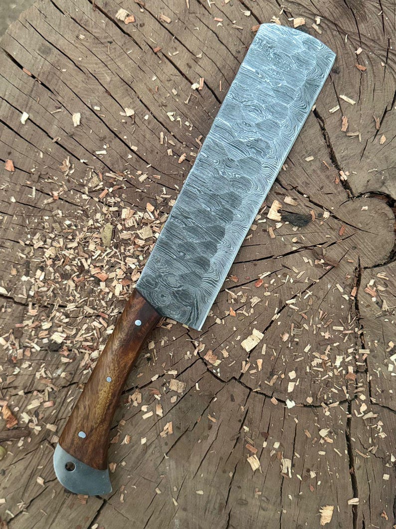 May include: A large, rectangular cleaver knife with a patterned steel blade and a wooden handle. The handle is secured with rivets and has a metal end cap. The knife rests on a wooden surface with wood shavings.