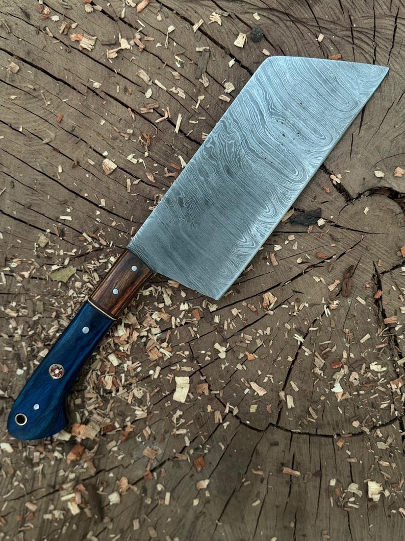 May include: A large, Damascus steel cleaver knife with a wooden and blue handle. The blade has a unique pattern and is set against a rustic wooden background. The handle is a combination of dark wood and blue, with decorative rivets.