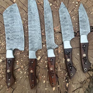 May include: A set of six Damascus steel knives with wooden handles. The blades have a unique patterned design. The handles are dark brown with decorative metal accents. The knives are arranged on a rough-hewn wooden surface.