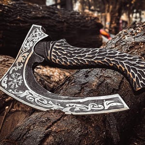 May include: A handcrafted axe with a dark, textured wooden handle and a silver-coloured blade. The blade features intricate engraved designs, including a symbol resembling a compass rose. The axe rests on a bed of dark brown logs.