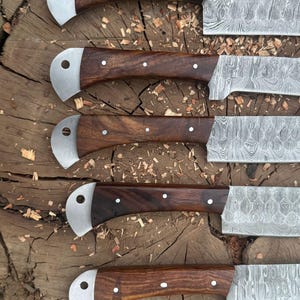 May include: Close-up of five Damascus steel knives with dark wood handles. The handles are secured with small white rivets and have a curved, silver-colored bolster. The blades display a unique, patterned design. The knives are arranged on a rough, wooden surface.