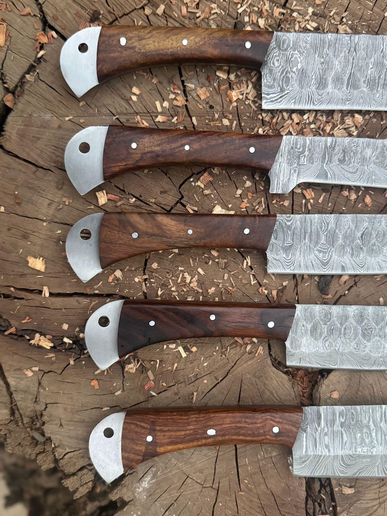 May include: Close-up of five Damascus steel knives with dark wood handles. The handles are secured with small white rivets and have a curved, silver-colored bolster. The blades display a unique, patterned design. The knives are arranged on a rough, wooden surface.