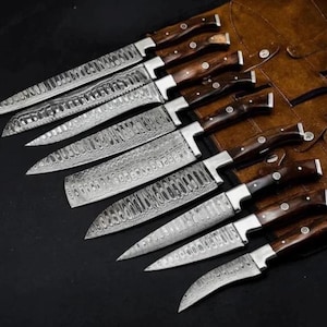 May include: A set of eight kitchen knives with patterned Damascus steel blades and dark brown wooden handles, arranged in a diagonal line. Each knife features a silver bolster and three rivets on the handle, displayed alongside a brown leather roll.