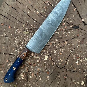 May include: A Damascus steel chef's knife with a dark blue handle and a wooden bolster. The blade has a unique swirling pattern. The knife is resting on a wooden surface with wood shavings scattered around it.