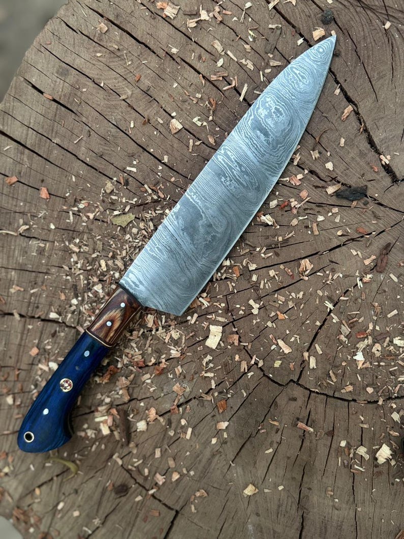 May include: A Damascus steel chef's knife with a dark blue handle and a wooden bolster. The blade has a unique swirling pattern. The knife is resting on a wooden surface with wood shavings scattered around it.