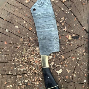 May include: A Damascus steel cleaver knife with a black handle and brass bolster. The blade has a unique pattern and a hole near the top. The handle is curved and has a polished finish. The knife is resting on a wooden surface with wood shavings.