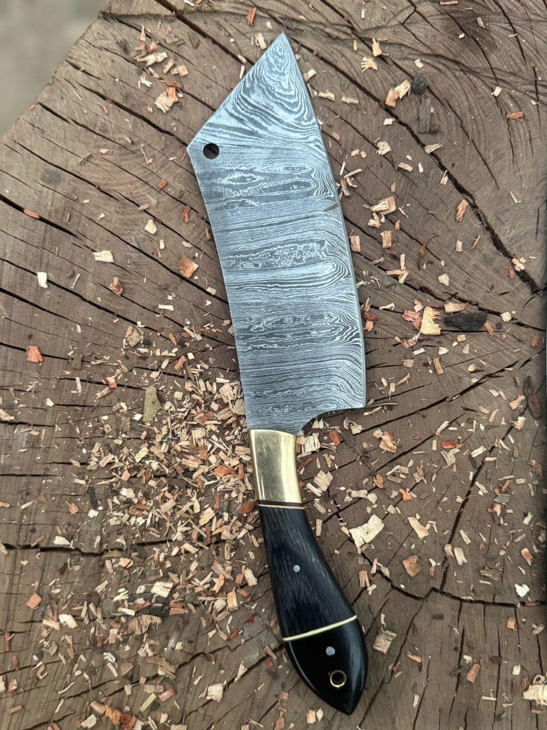 May include: A Damascus steel cleaver knife with a black handle and brass bolster. The blade has a unique pattern and a hole near the top. The handle is curved and has a polished finish. The knife is resting on a wooden surface with wood shavings.