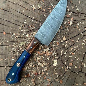 May include: A handcrafted chef's knife with a patterned steel blade and a handle featuring blue, brown, and copper accents. The handle has decorative rivets. The knife is designed for kitchen use.
