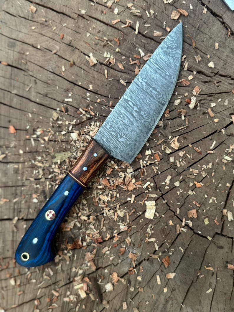 May include: A handcrafted chef's knife with a patterned steel blade and a handle featuring blue, brown, and copper accents. The handle has decorative rivets. The knife is designed for kitchen use.