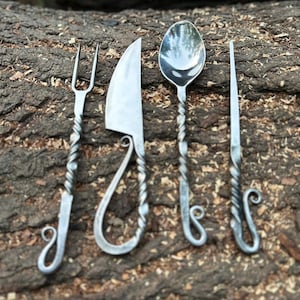 May include: Hand-forged metal flatware set including a fork, knife, spoon, and a slender utensil. The handles have a twisted design and end in a decorative curl. The set is displayed on a textured, brown surface.