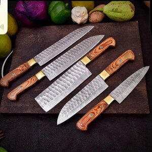 May include: A collection of six kitchen knives with wooden handles and patterned blades. The knives are arranged on a dark wooden cutting board. The handles are a light brown color with brass bolsters. The blades have a textured, hammered appearance.