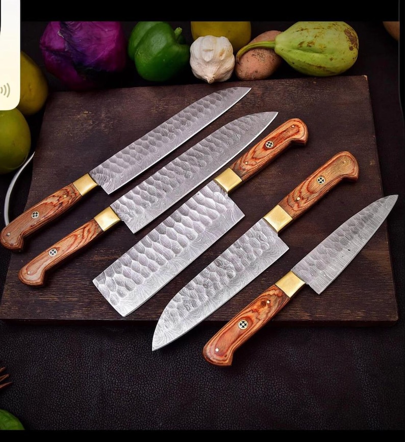 May include: A collection of six kitchen knives with wooden handles and patterned blades. The knives are arranged on a dark wooden cutting board. The handles are a light brown color with brass bolsters. The blades have a textured, hammered appearance.